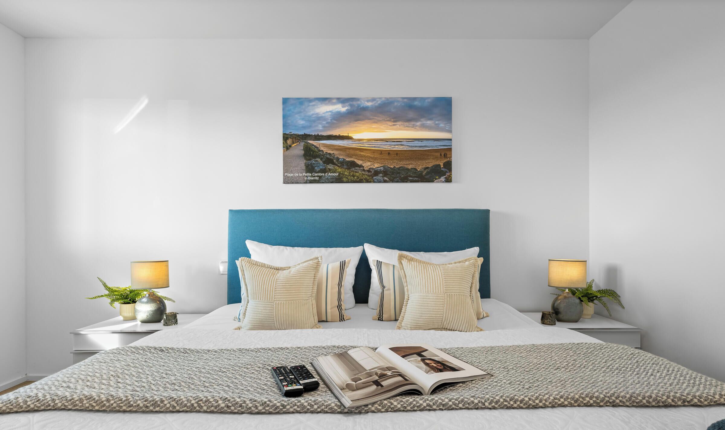 Ein Schlafzimmer mit einem Bett, Kissen, einer Fernbedienung, einer Zeitschrift und einem Gemälde mit einer Strandansicht.