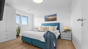 Un dormitorio con cama, mesitas de noche, ventana, un cuadro y una puerta.