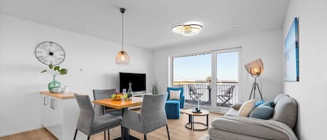 A modern apartment with a dining area, a living room with a sofa, a balcony with a view, and a large clock on the wall.