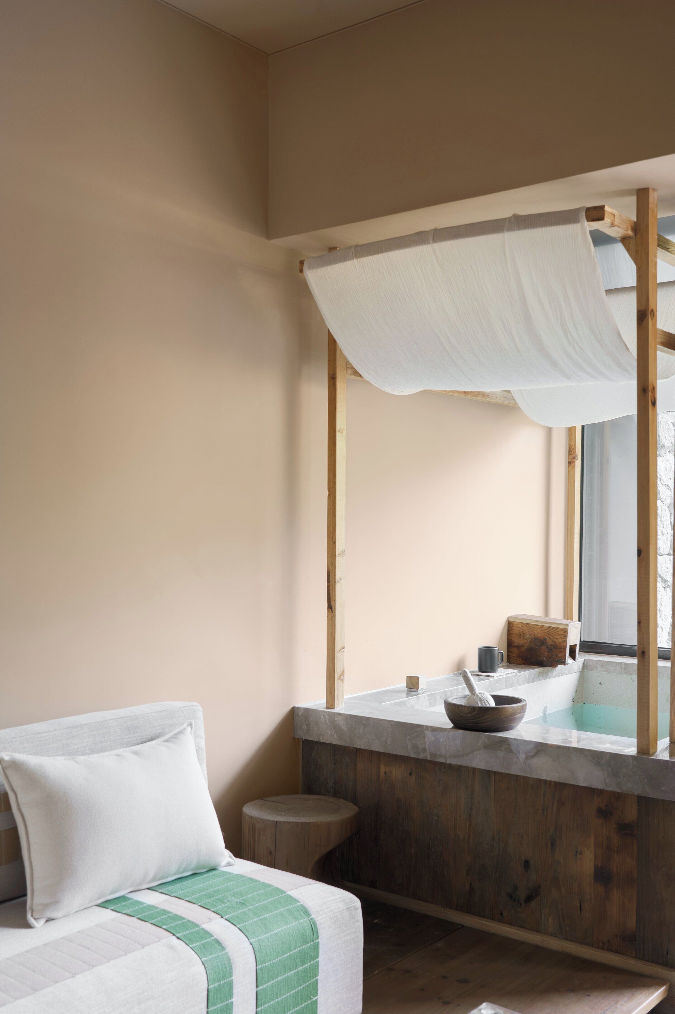 A room with a bed, a wooden stool, a small table with a bowl, and a window with a view.