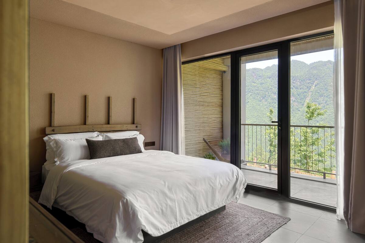 Ein Schlafzimmer mit einem großen Bett, Blick auf Grün hinter einer Schiebetür und einem Balkon.