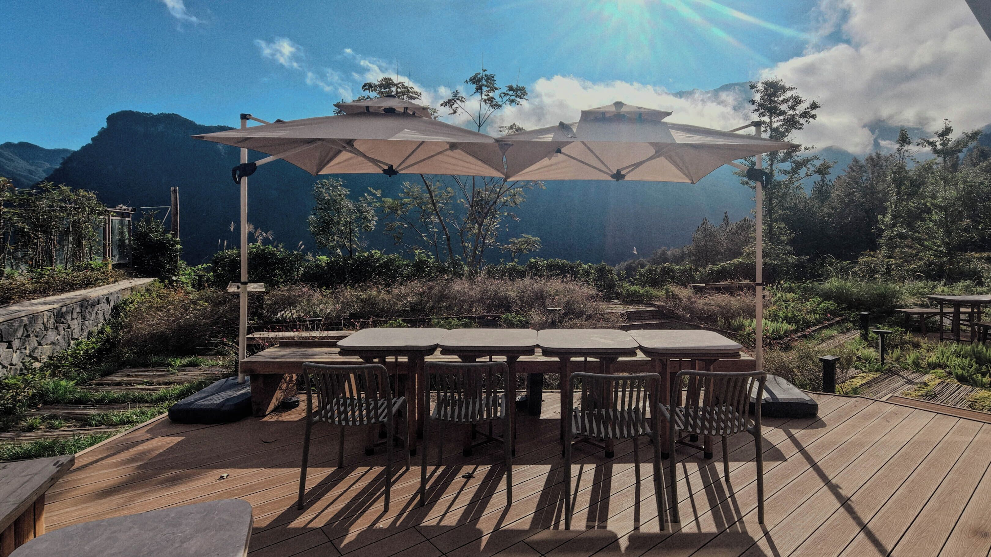 An outdoor dining area with a wooden deck, tables, chairs, and large umbrellas providing shade.