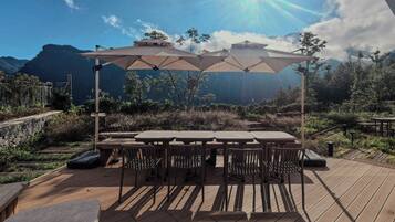 An outdoor dining area with a wooden deck, tables, chairs, and large umbrellas providing shade.