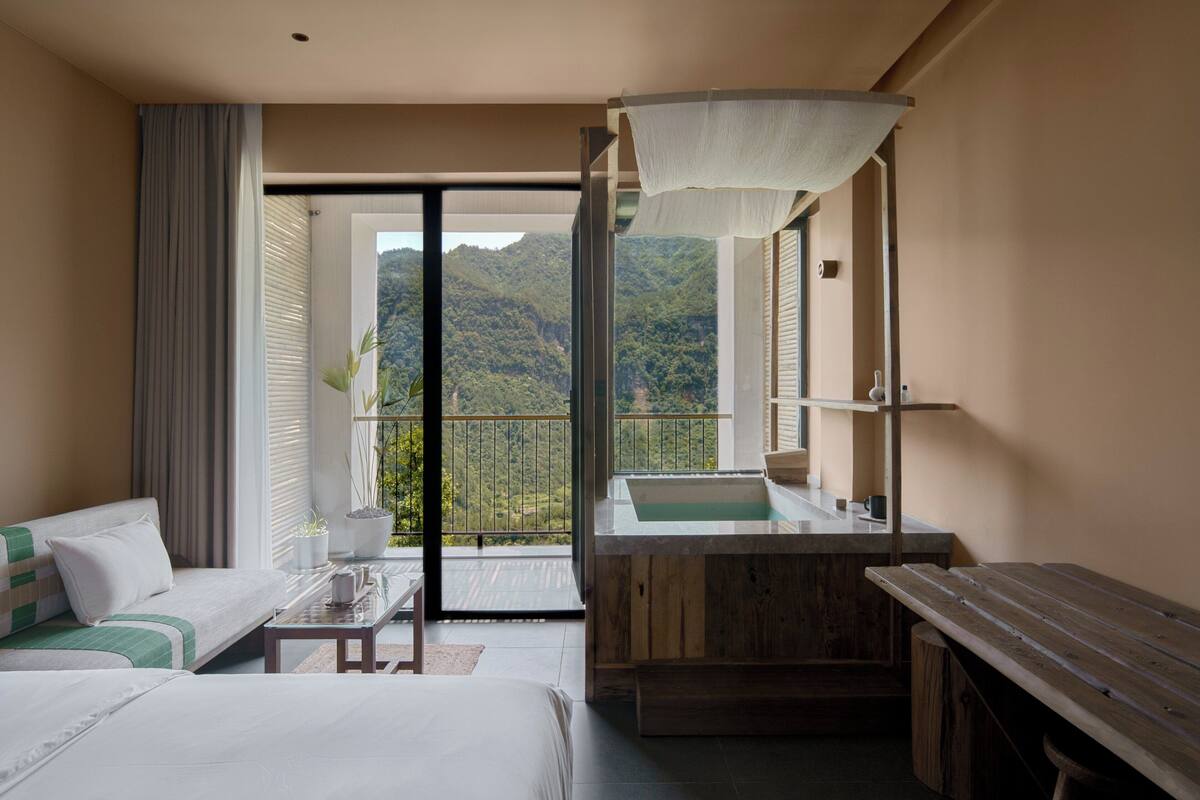 Ein Hotelzimmer mit einem großen Bett, einem Holztisch, einem kleinen Tischchen, einem Waschbecken und einem Balkon mit Bergblick.