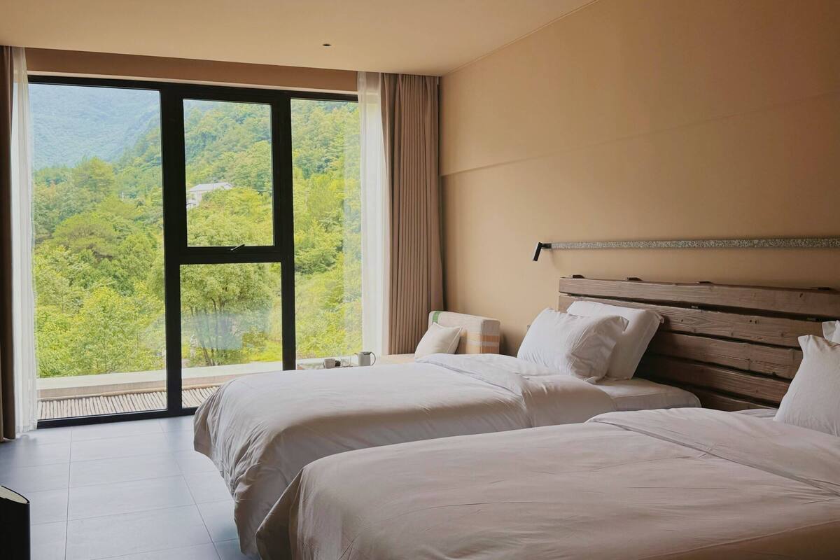 Ein Hotelzimmer mit zwei Betten, einem großen Fenster mit Blick auf eine bewaldete Landschaft und einem hölzernen Kopfteil.