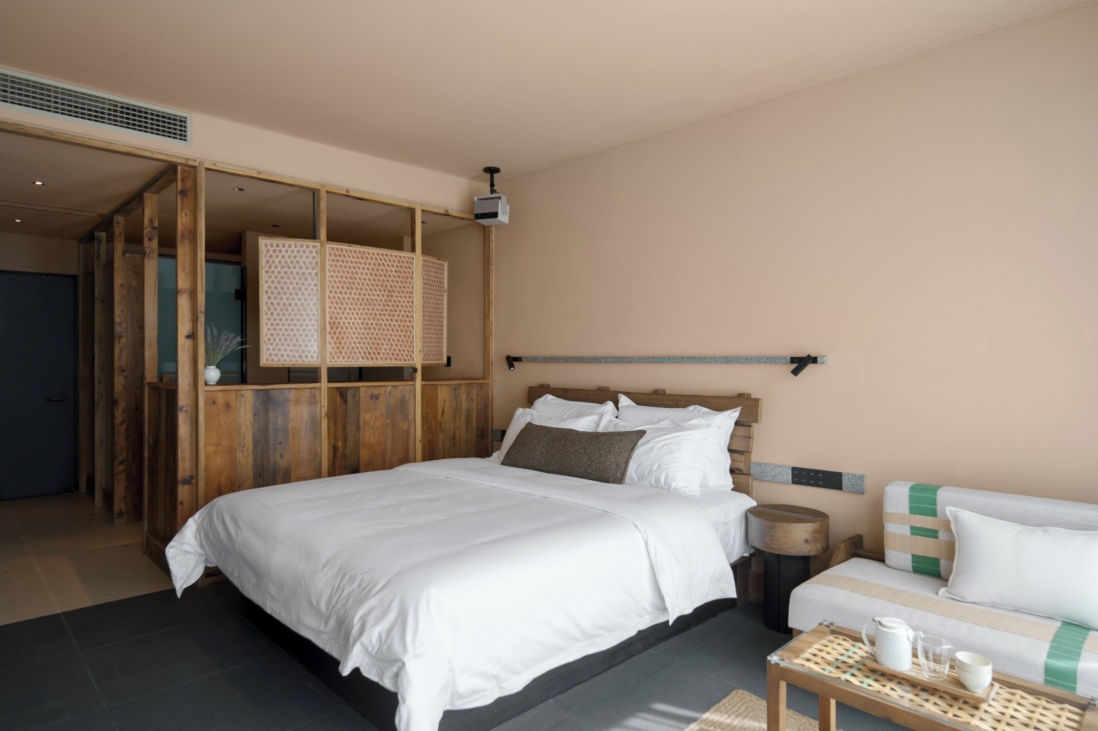 Habitación de hotel moderna con una cama grande, un panel de madera, una mesita con jarrón y taza, y un sofá con cojines.