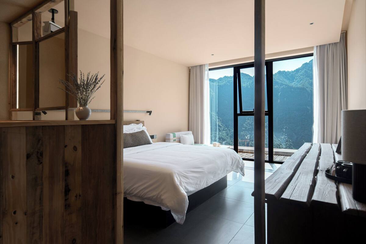 Ein modernes Hotelzimmer mit einem großen Bett, einem Holztisch und Blick auf Berge durch ein großes Fenster.