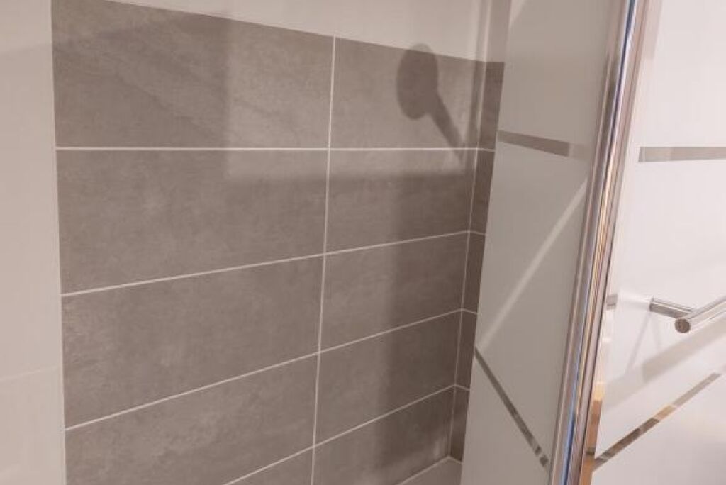 A bathroom with grey tiled walls and a glass shower enclosure.