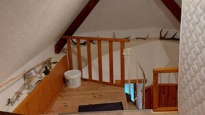 Un petit espace mansardé aux poutres apparentes, un petit escalier en bois et un seau blanc posé sur le sol.
