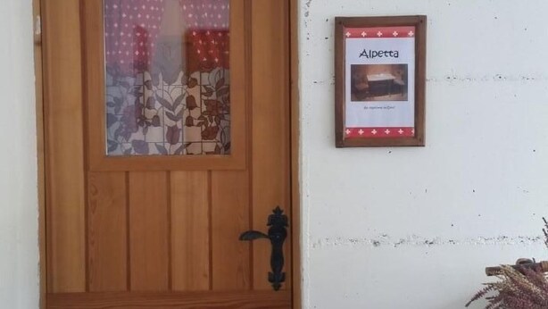 Una puerta de madera con un panel de vidrio, un pequeño cuadro enmarcado con la etiqueta "Alpetta" en la pared, y una cesta de mimbre a la derecha.