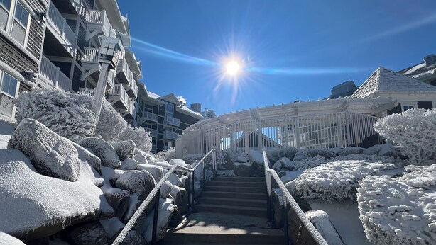 Eine schneebedeckte Treppe führt zu einem Gebäude mit Balkonen, umgeben von schneebeschichteten Büschen und Felsen.