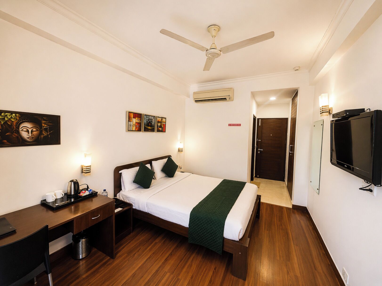 A hotel room with a bed, desk, chair, television, artwork, and a ceiling fan.