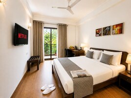 Habitación de hotel con cama, escritorio, silla, televisor, balcón con cortinas y cuadros en la pared.