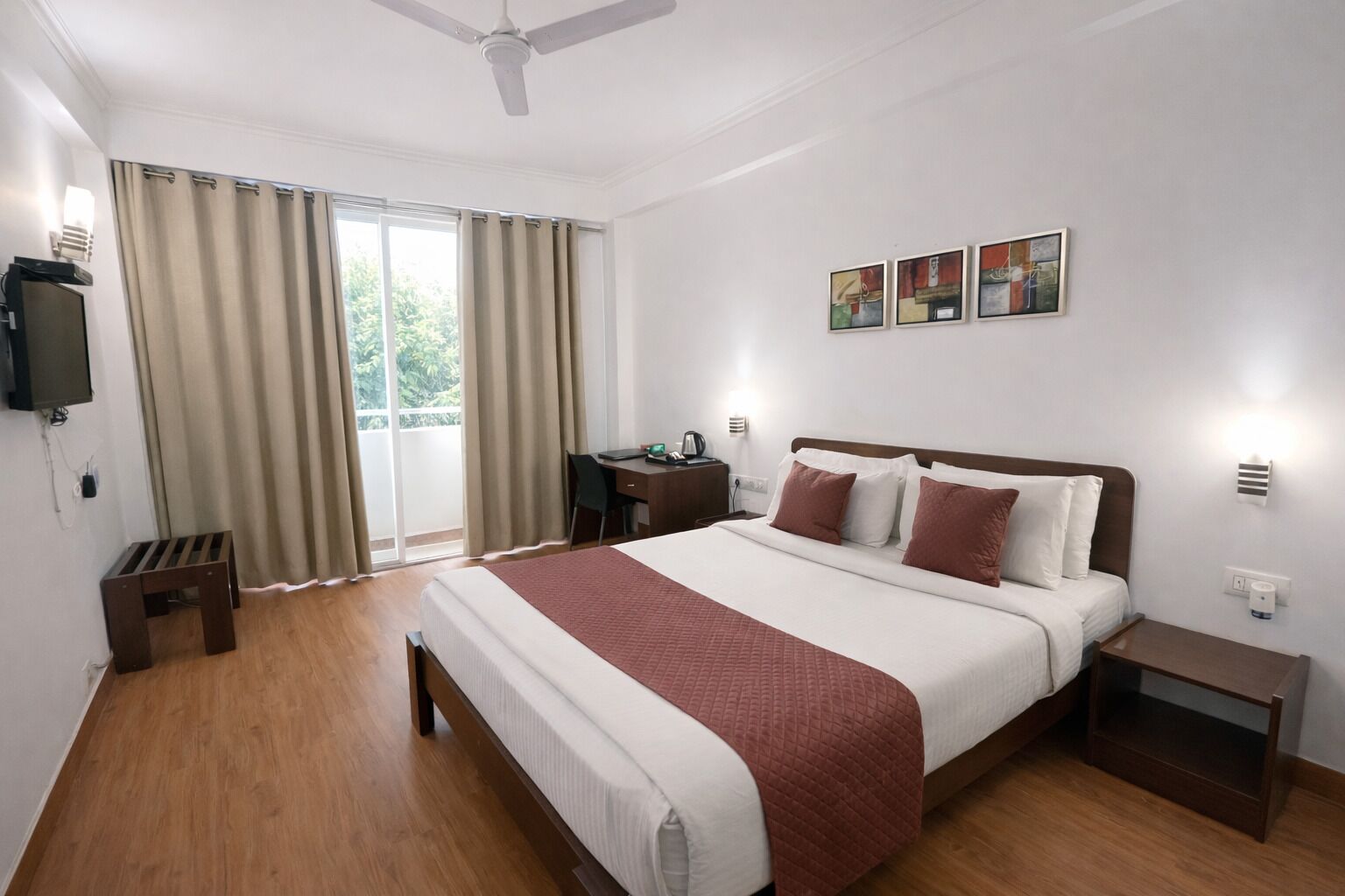 A hotel room with a bed, bedside tables, a desk, a chair, a television, and a window with curtains.