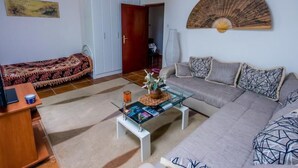 Een woonkamer met een bank, een glazen salontafel, een bed met een bedsprei met patroon, een houten kast en een ventilator aan de muur.