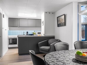 A modern kitchen with grey cabinets, a microwave, and a sink. There is a dining area with a round table and chairs, and a window with a view of trees. - "Allyson" -  from the sea by Interhome (Hadsund)