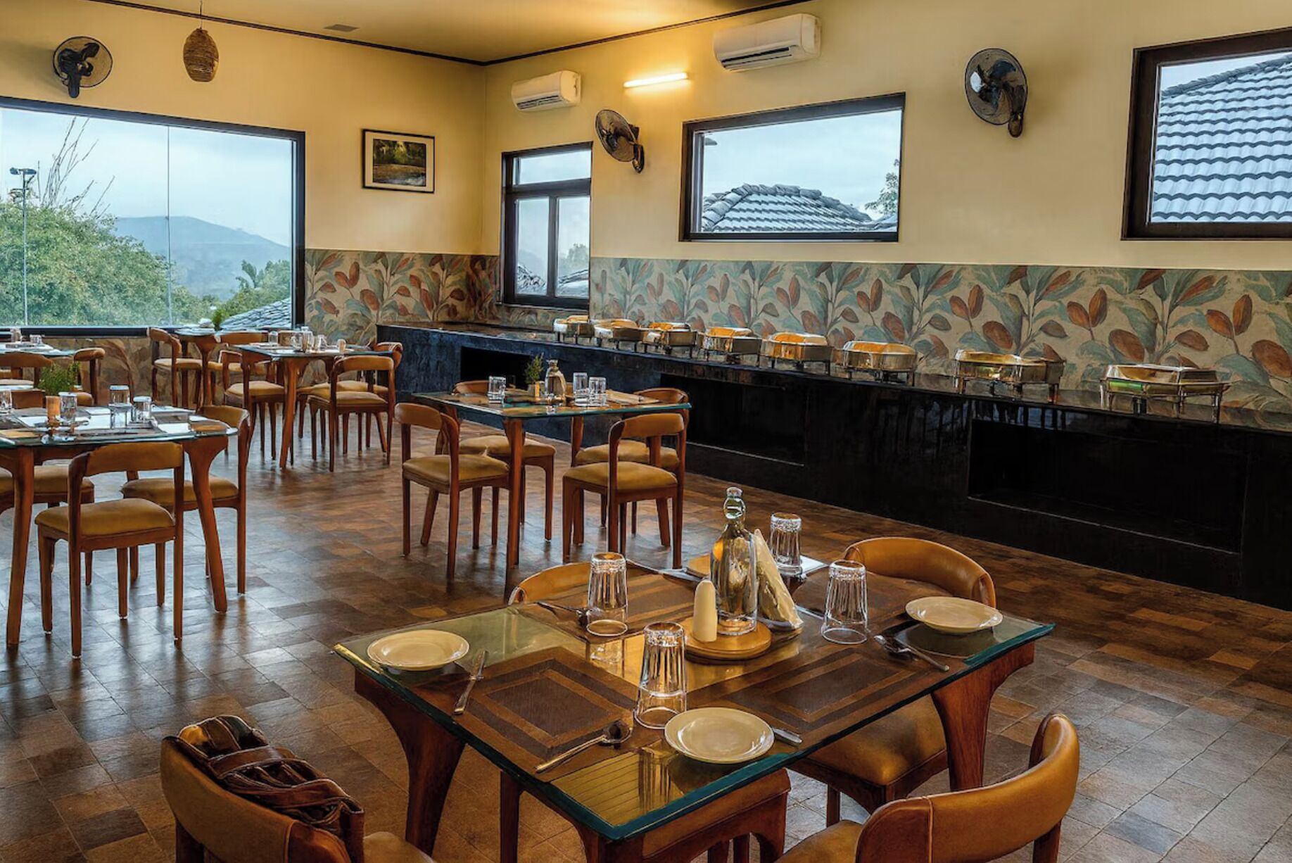 A restaurant with wooden tables and chairs, a bar area, and large windows offering a view of the outdoors.