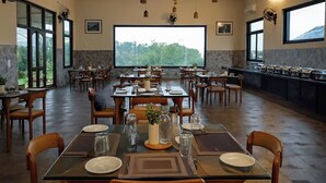 A restaurant with multiple tables set for dining, large windows offering a view of the outdoors, and a stone wall on one side. - Sonemale Resort Chikmagalur (Mudigere)