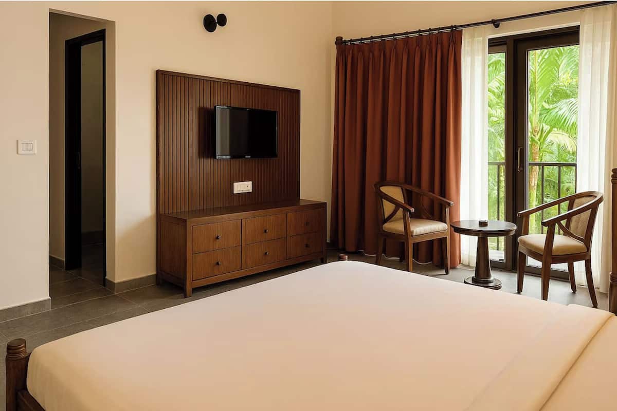 A hotel room with a large bed, a TV mounted on a wooden cabinet, two chairs, and a view of greenery through a window with brown curtains.