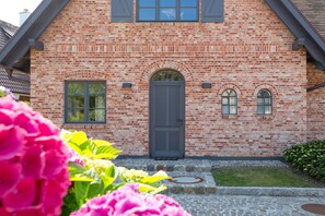 Une maison en briques avec une porte grise, deux fenêtres et un petit jardin où poussent des fleurs roses.