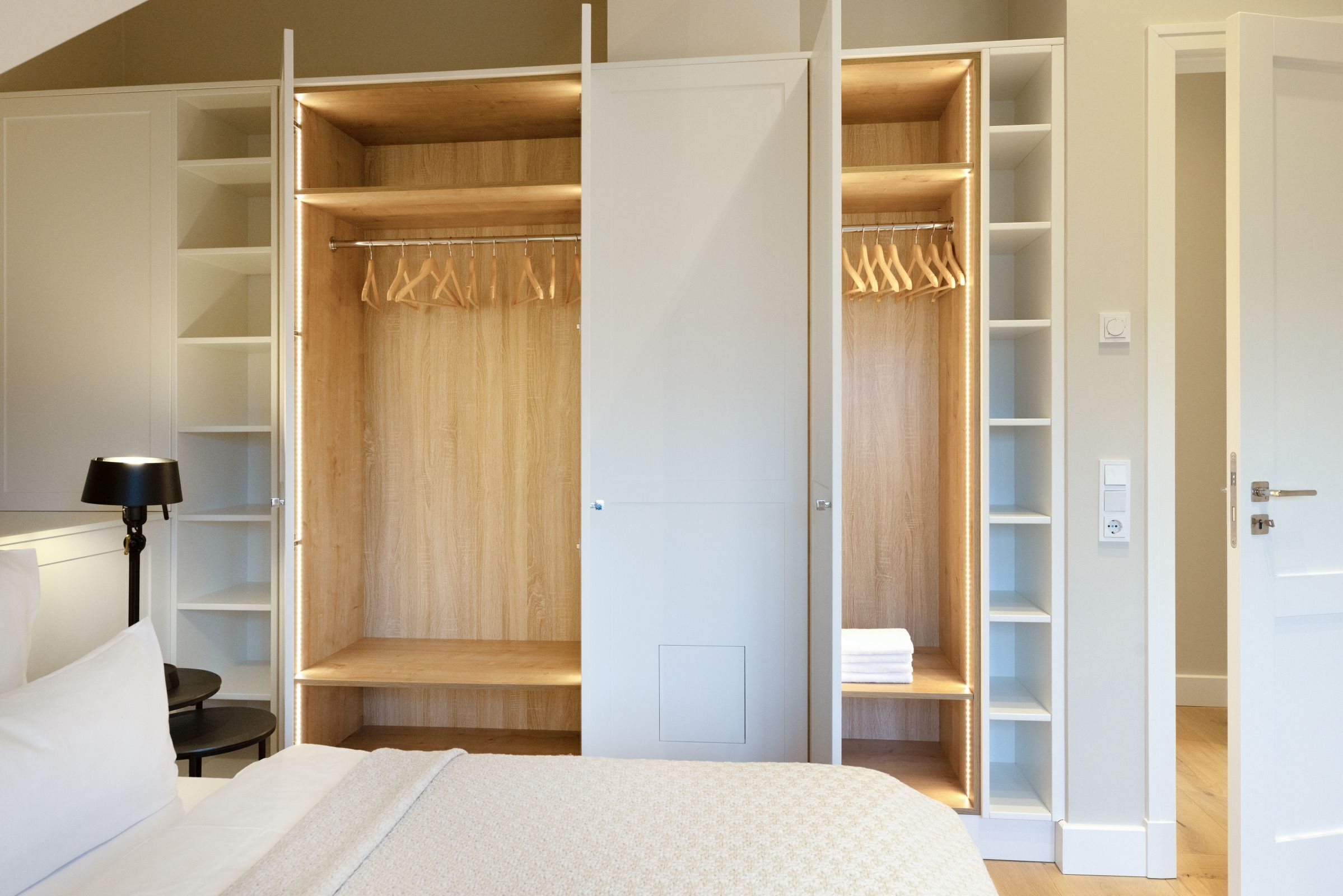 Ein Schlafzimmer mit großem begehbaren Kleiderschrank, einem Bett mit weißer Bettwäsche, einem Nachttisch mit Lampe und Holzboden.