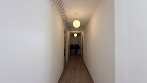 A narrow hallway with a wooden floor, white walls, and a single hanging light fixture. - Brown H (Bangkok)