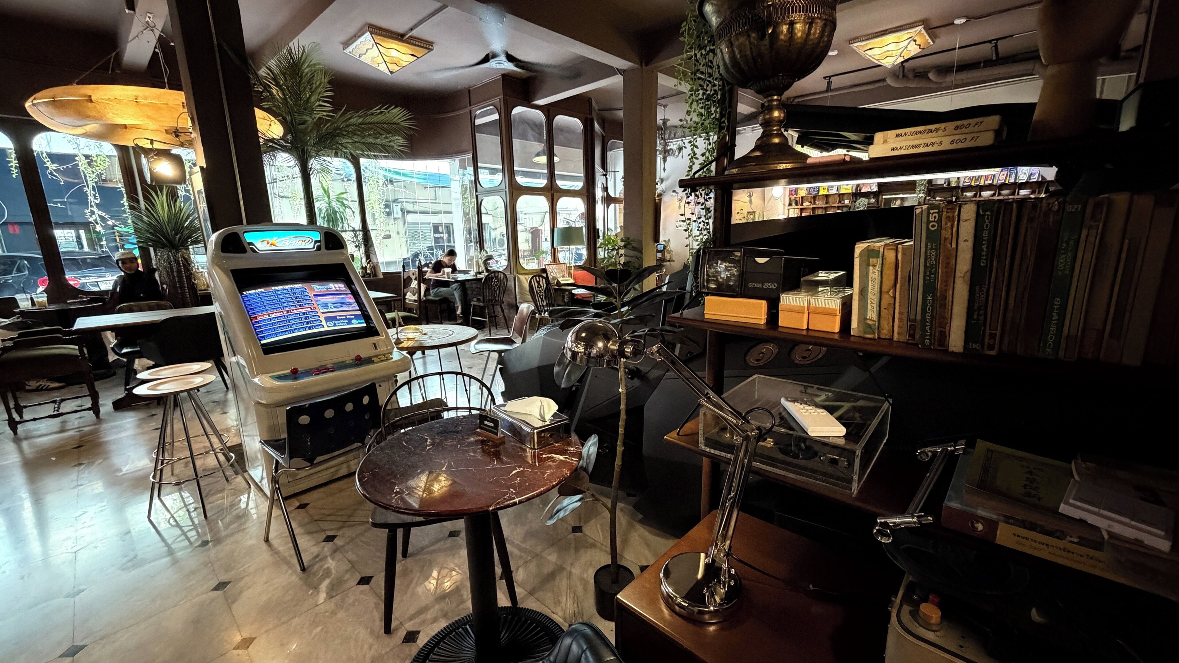 A room with a large window, a jukebox, a table with a chair, and a shelf with books and decorative items.