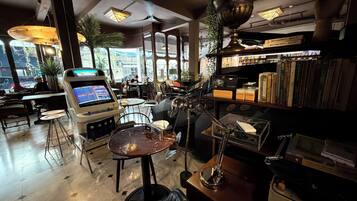 A room with a large window, a jukebox, a table with a chair, and a shelf with books and decorative items.