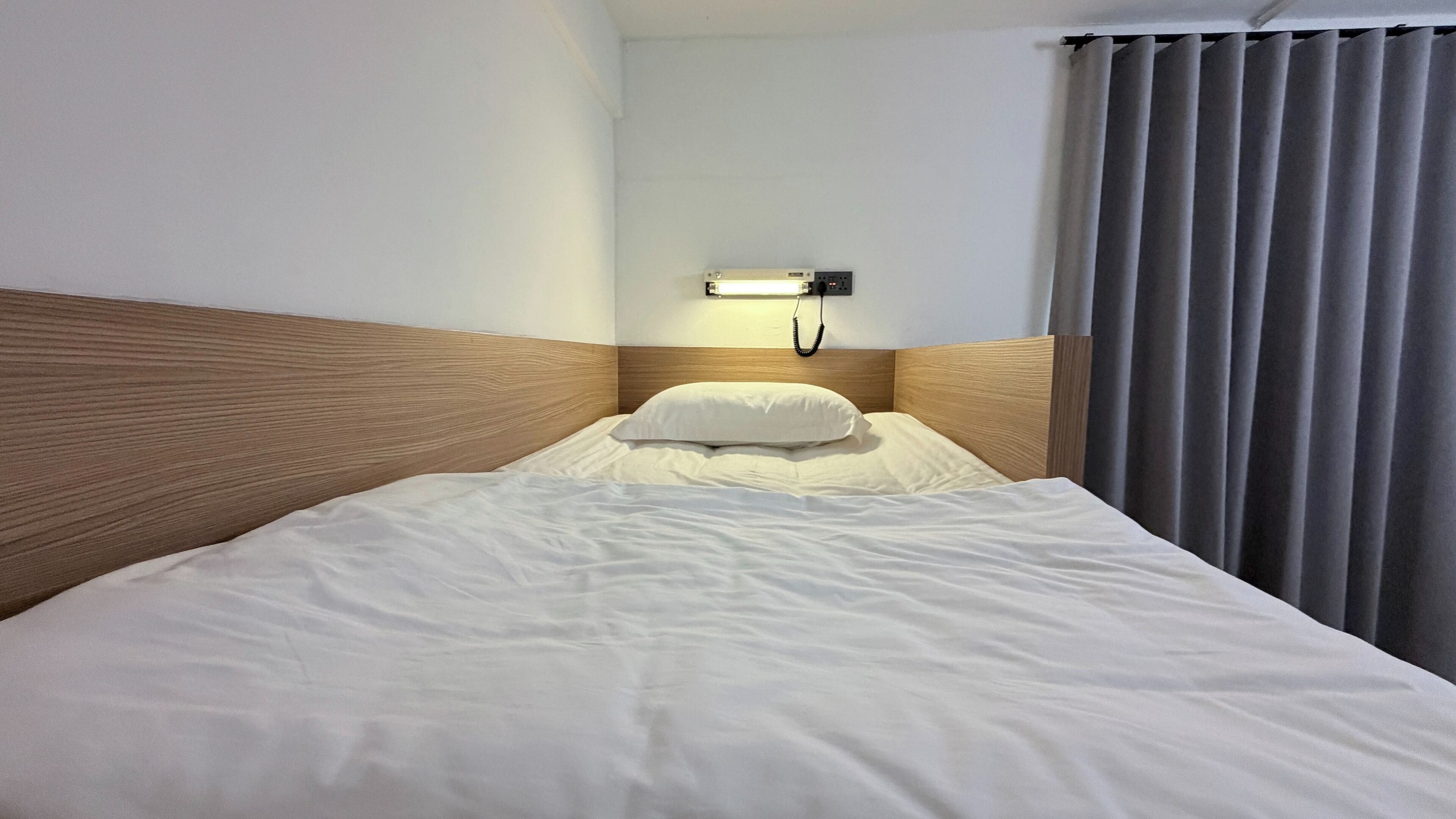 Une chambre d’hôtel avec une tête de lit en bois, un lit blanc avec un oreiller, un téléphone sur la table de chevet et une fenêtre avec des rideaux gris.