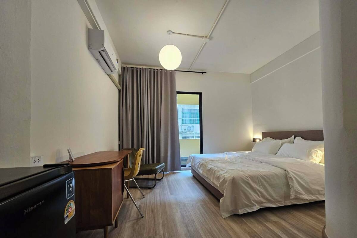 A hotel room with a bed, a desk, and a chair. There is a window with curtains, an air conditioning unit, and a ceiling light.