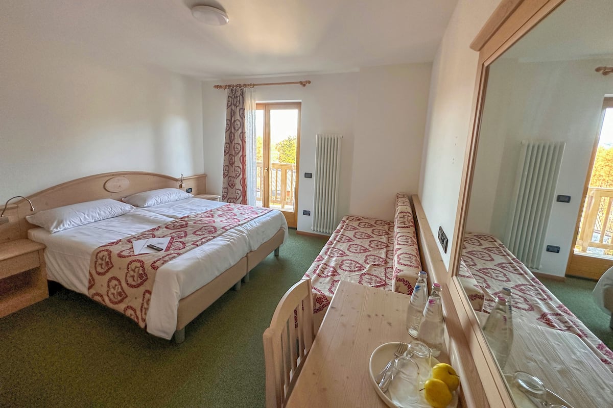 A hotel room with a double bed, a wooden nightstand, a dresser with a mirror, and a balcony door.
