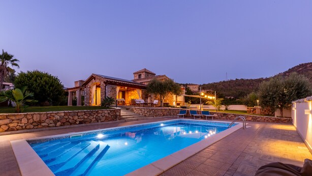 Una piscina con pared de piedra y sillas de descanso, rodeada por un patio de piedra y un edificio con fachada de piedra.
