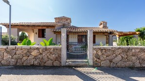 Un muro de piedra con una reja de metal, una casa con techo de tejas y un cielo azul despejado.