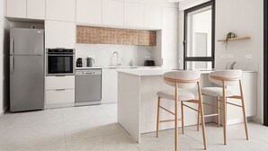 Cocina moderna con refrigerador, horno y lavavajillas de acero inoxidable, una isla blanca con taburetes y una ventana con persianas.