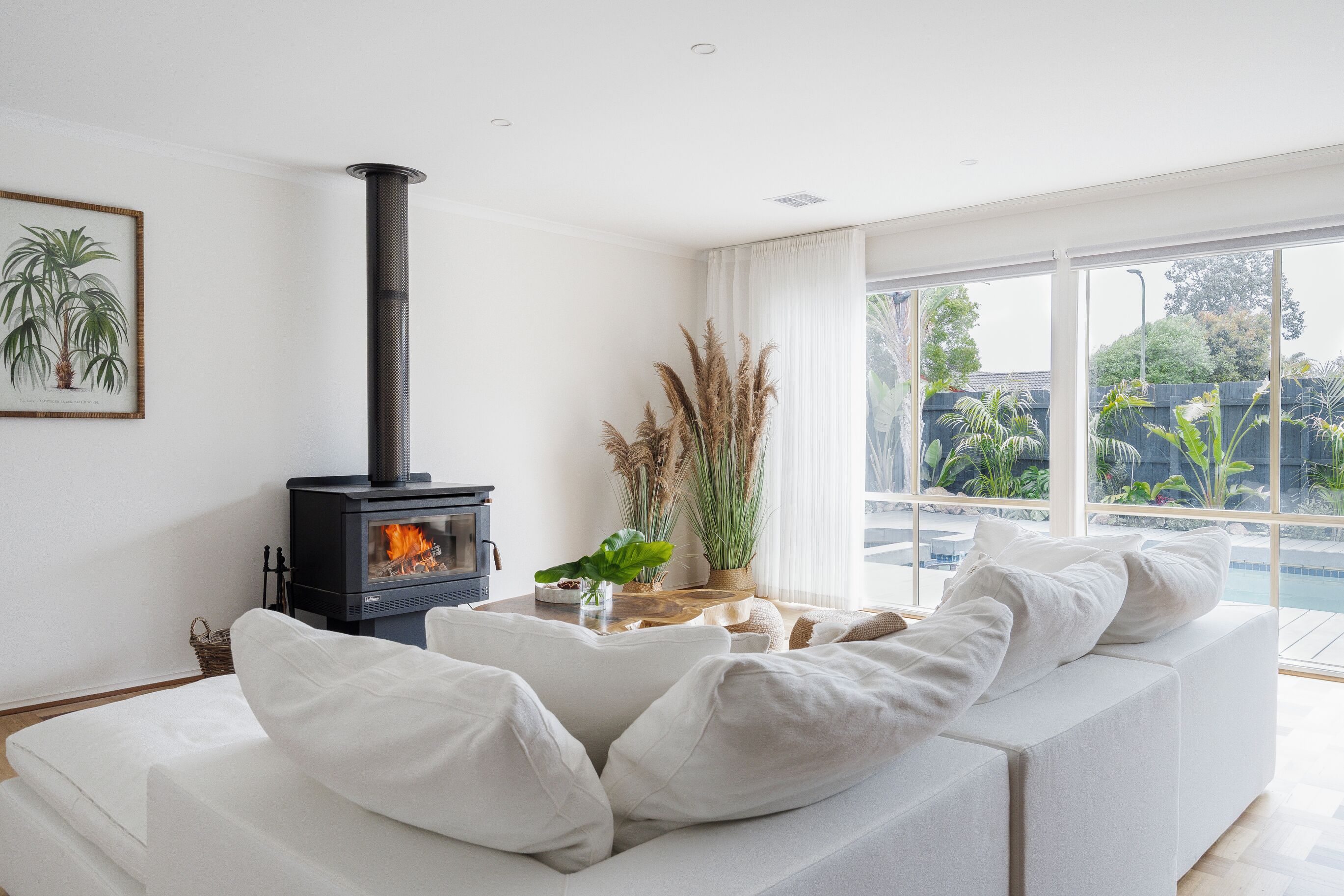 Un salon moderne avec un canapé blanc, un poêle à bois noir, une grande fenêtre donnant sur un paysage verdoyant et une illustration botanique encadrée accrochée au mur.