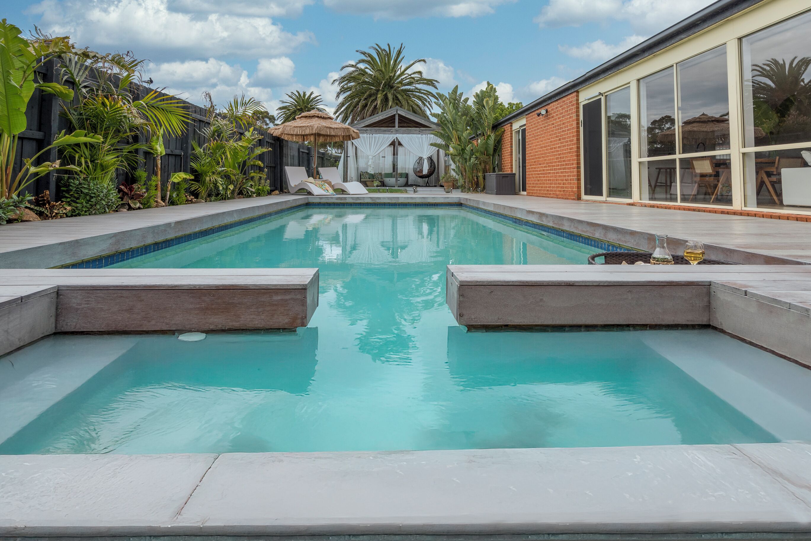 Une piscine dotée d’une structure à toit de chaume, entourée d’une végétation tropicale luxuriante et d’un bâtiment moderne avec de grandes fenêtres.