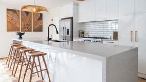 A modern kitchen with a large island, white cabinetry, stainless steel appliances, and wooden bar stools. - Bahagia Oasis - 4pm check out Sundays* (Safety Beach)