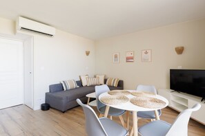 Ein modernes Wohnzimmer mit einem grauen Sofa, einem runden Esstisch mit gewobenen Untersetzern und einem Flachbildfernseher auf einem weißen Schrank.
