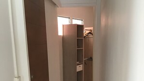 Ein schmaler Flur mit einer Holztür links, einem Schrank mit Regalen und Kleiderstange rechts sowie einem gefliesten Boden.