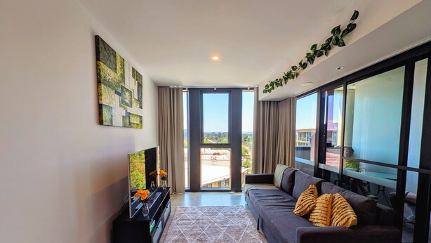 A modern living room with a sofa, a TV, and a large window offering a city view. - Centrally located new apartment with free parking and access of pool , gym (Canberra)