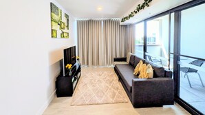 A modern living room with a sofa, a coffee table, and a TV. There is a large window with curtains and a balcony visible through the window. - Centrally located new apartment with free parking and access of pool , gym (Canberra)