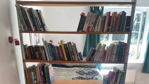A red sofa with colorful cushions, a wooden bookshelf filled with books, and a window with curtains. - Artistic, 1930's studio-flat - 20 mins to Central London. (London)