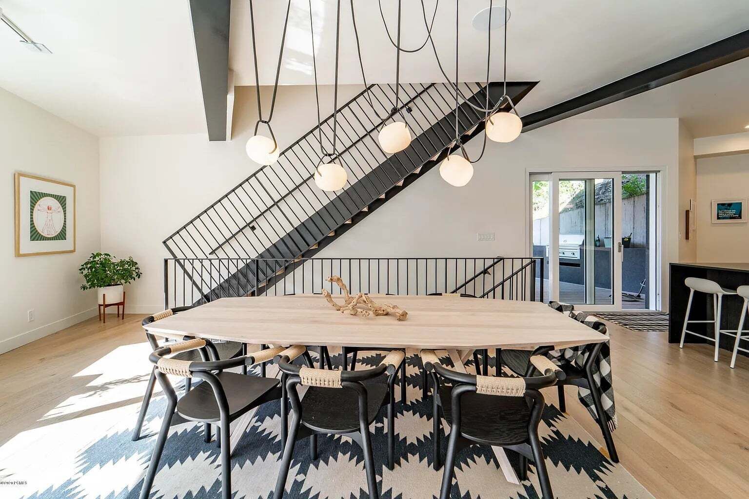 Un intérieur moderne avec une grande table à manger en bois au centre, des chaises noires, un escalier doté d’une rampe en métal et des luminaires suspendus.
