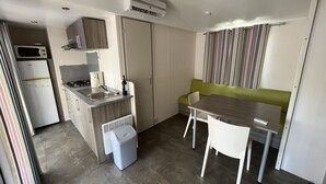 Een kleine keuken met een magnetron, kookplaat, spoelbak en een kleine prullenbak. Er is een zithoek met een tafel en stoelen, en een groene bank.