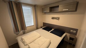 Een netjes opgemaakt bed met kussens en een opgevouwen plaid, een nachtkastje met een lamp, en een raam met gordijnen.