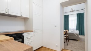 A compact kitchen with white cabinets, a wooden countertop, and a built-in oven. A hallway leads to a bedroom with a bed, a chair, and a window with curtains. - RentPlanet - Apartament Chmielna (Warsaw)