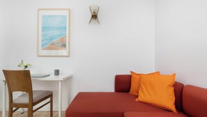 A room with a wooden chair, a white desk, a red sofa, and a framed picture of a beach. - Cozy Studio in the Center | Workspace (Gdynia)