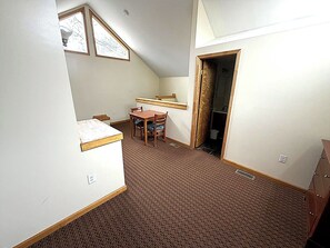 Ein kleiner, kompakter Raum mit schrägen Wänden, einem Esstisch mit Stühlen, einem kleinen Fenster und einer Tür zu einem Badezimmer.
