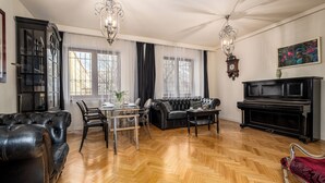 Un salón amplio con suelo de parquet de madera, un sofá de cuero negro, una mesa de comedor con sillas, un piano negro y una araña.