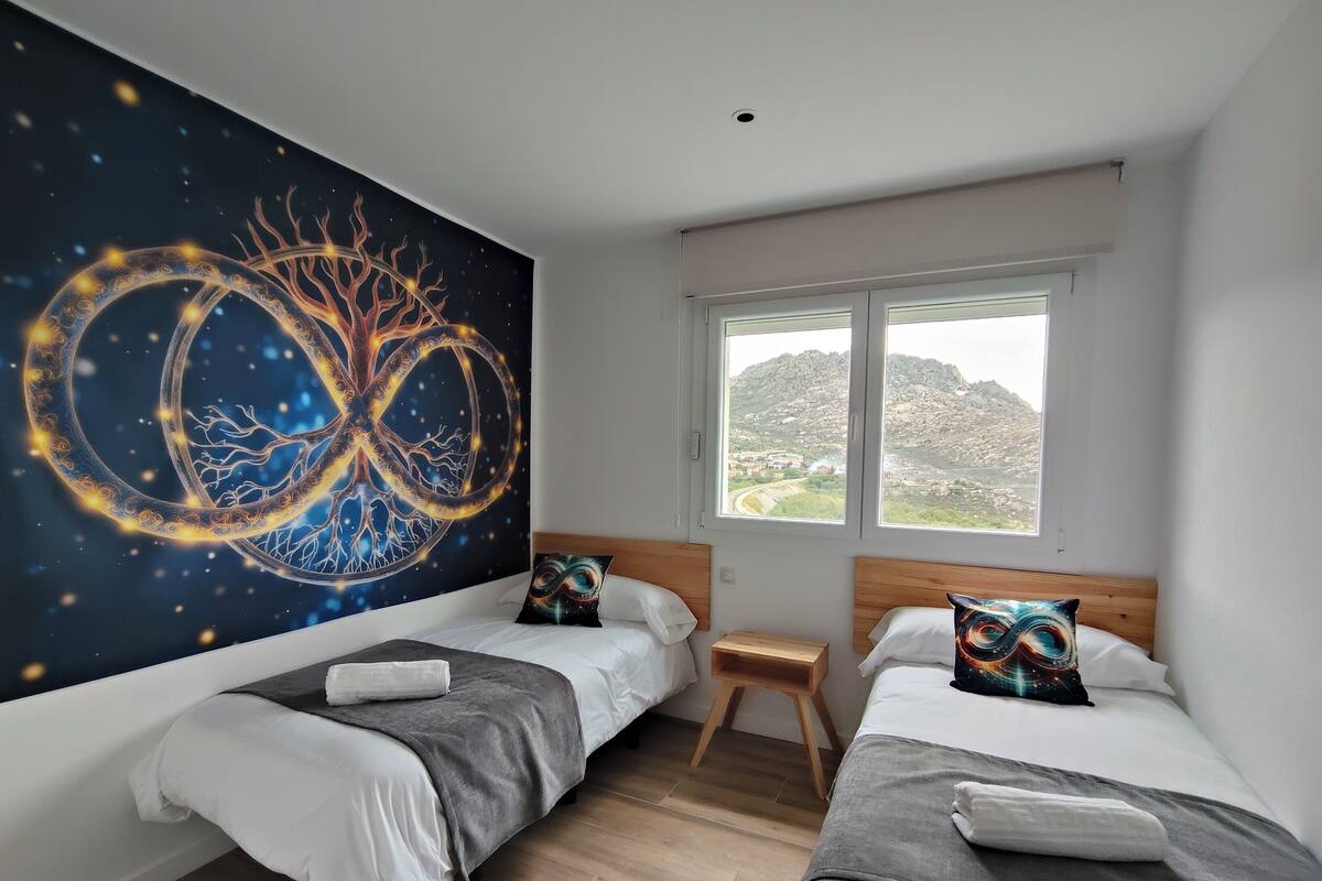 A bedroom with two beds, a large wall mural, a window with a view of a mountainous landscape, and a small wooden stool.